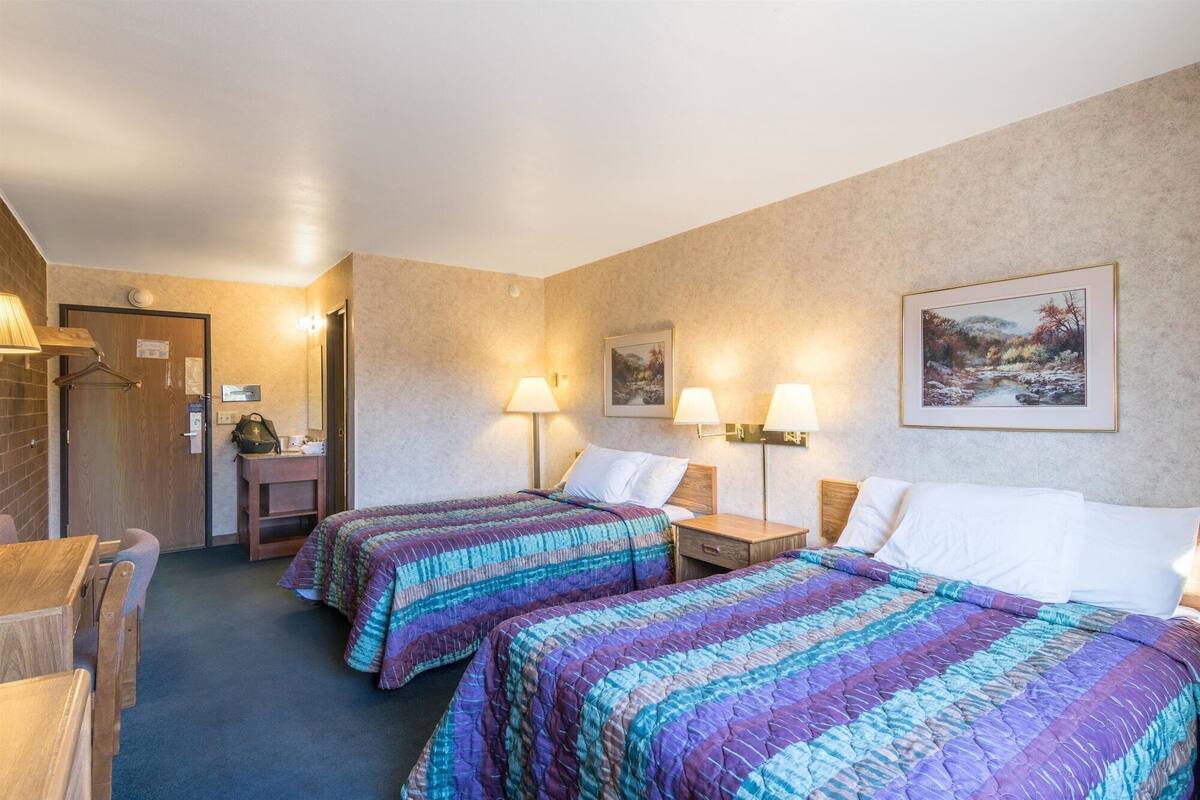 Een hotelkamer met twee bedden, een bureau, een stoel, een lamp en een schilderij aan de muur.