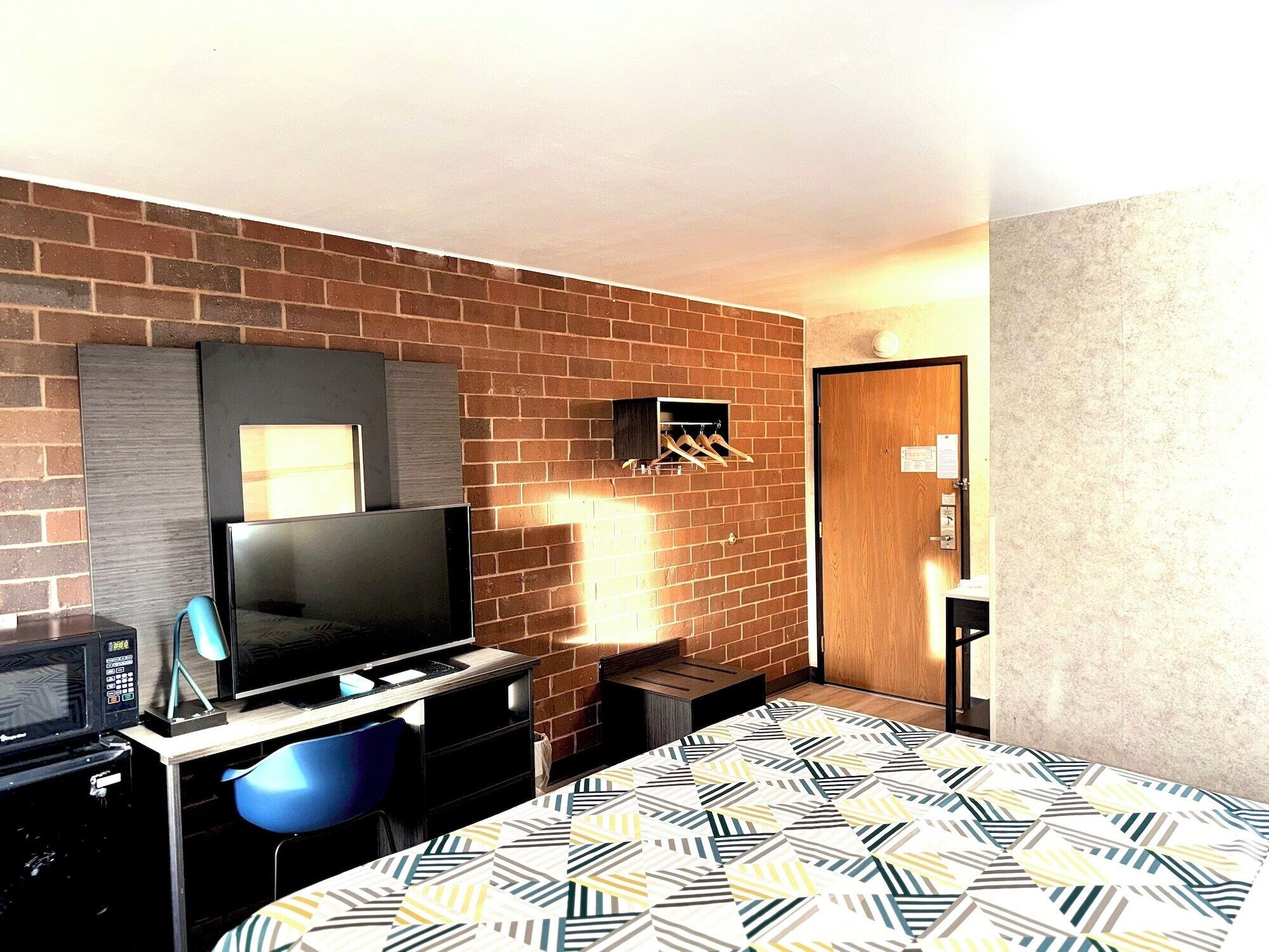 A hotel room with a brick wall, a flat-screen TV, a desk with a microwave, a blue chair, and a patterned bedspread.