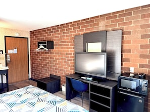A compact hotel room with a brick wall, a wooden door, a bed with patterned bedding, a desk with a computer, a TV, and a microwave.