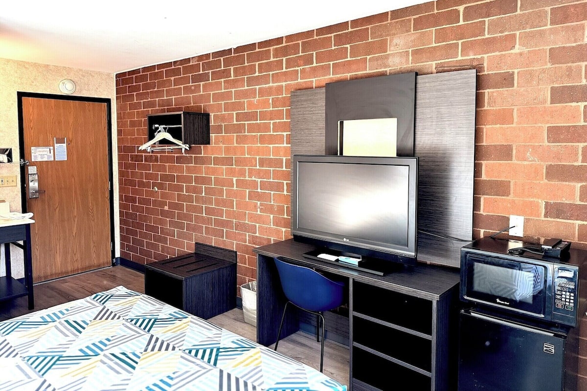 Een compacte hotelkamer met een bakstenen muur, een bed met beddengoed met een patroon, een bureau met een computer, een televisie en een magnetron.
