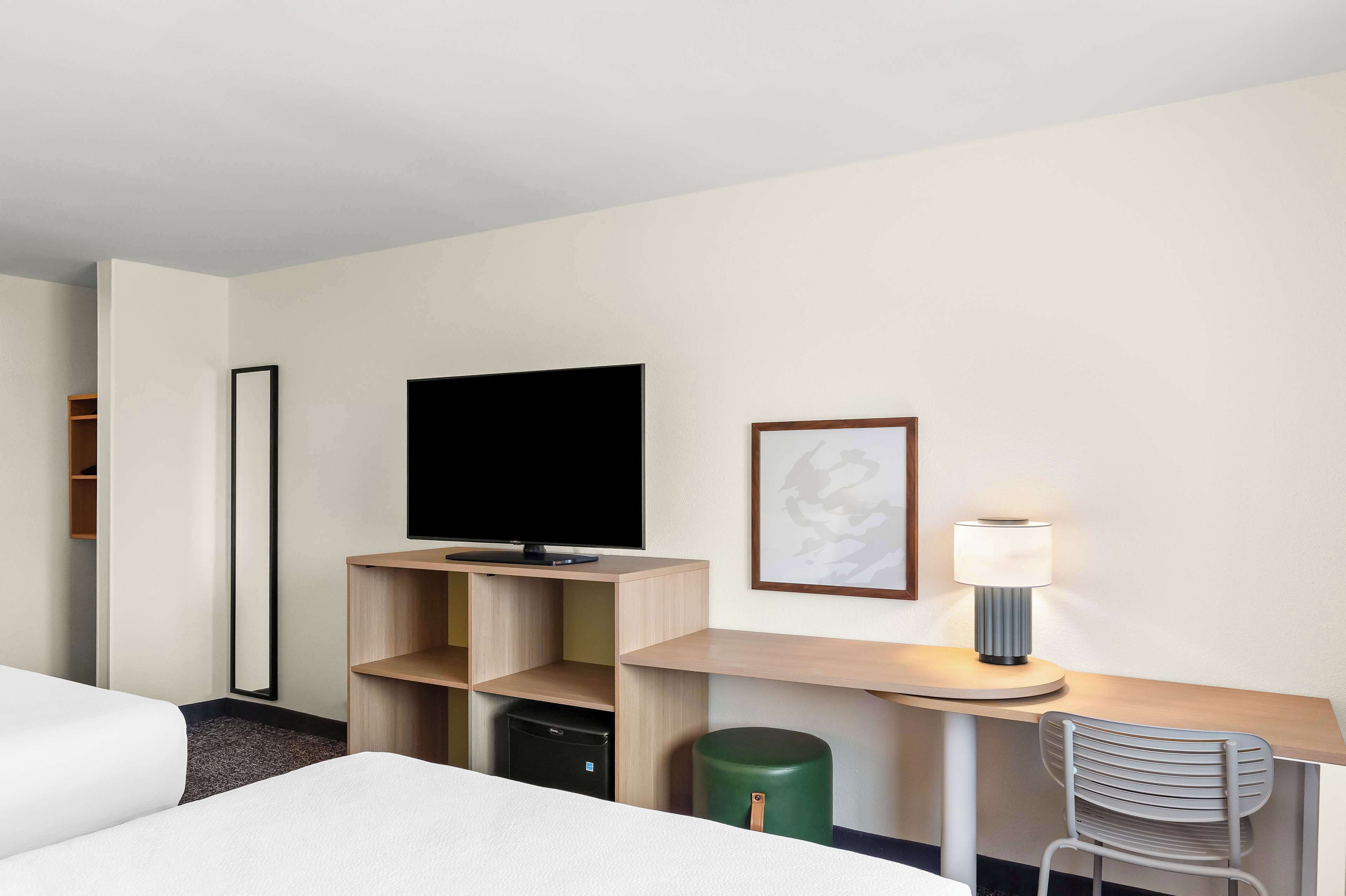 A hotel room with a bed, a desk with a lamp, a television, a trash can, and a chair.