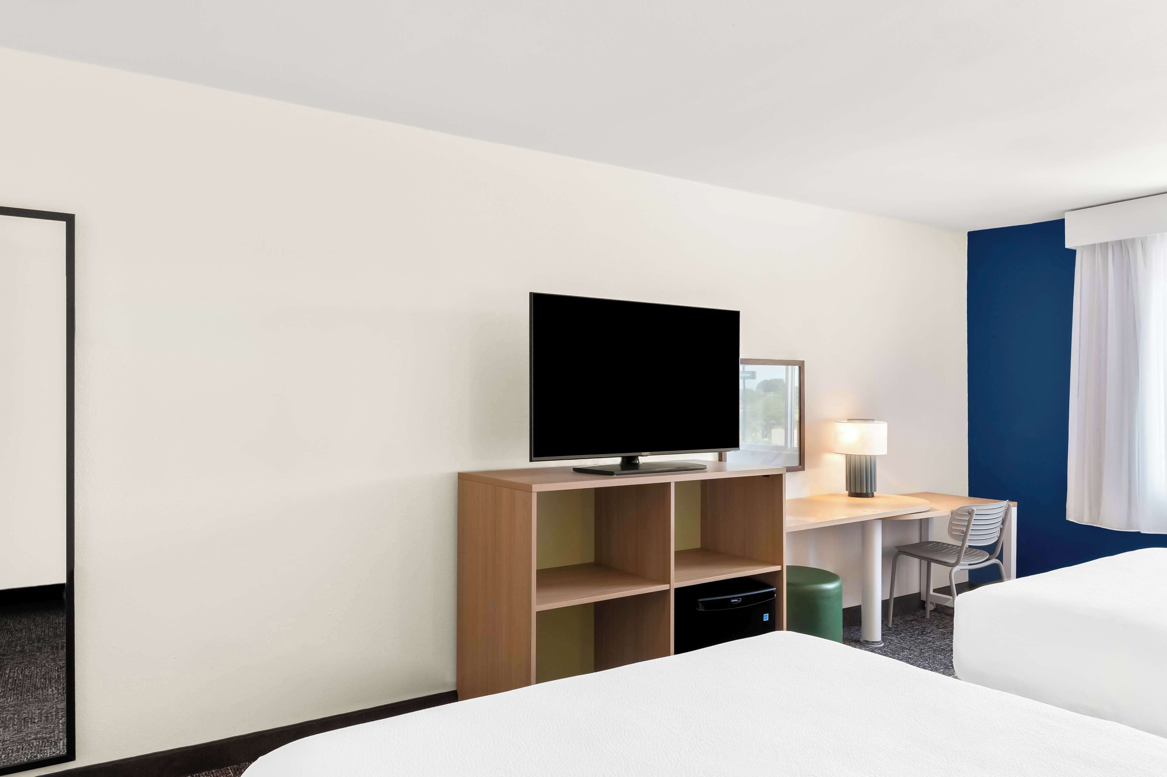 A hotel room with a bed, a desk with a flat-screen TV, a chair, a lamp, and a trash bin.
