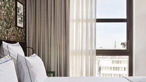 Ein Hotelzimmer mit Bett, weißen Leinentüchern, Blick auf den Eiffelturm und einem Fenster mit durchsichtigen Vorhängen.