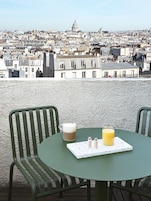 A terrace with a table and chairs, a view of a cityscape, and a table setting with a mug and a glass of orange juice.