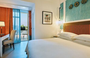 Ein Hotelzimmer mit Bett, Schreibtisch, Stuhl, einem Balkon mit Blick und einer Wand mit dekorativen Tellern.