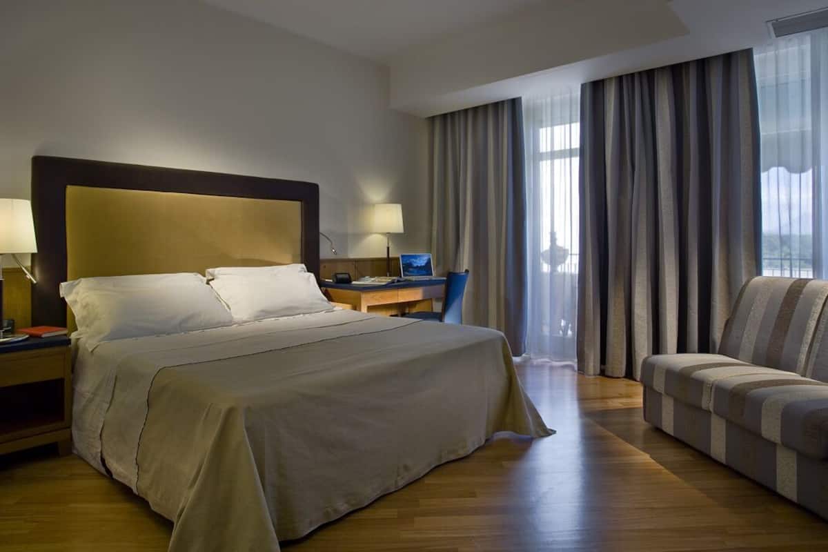 Une chambre d’hôtel avec un grand lit, un bureau équipé d’un ordinateur, une chaise et une vue sur la ville grâce à de grandes fenêtres.