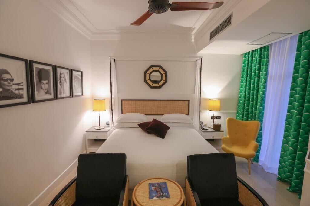 A hotel room with a bed, two bedside tables with lamps, a chair, a round table, and a ceiling fan.