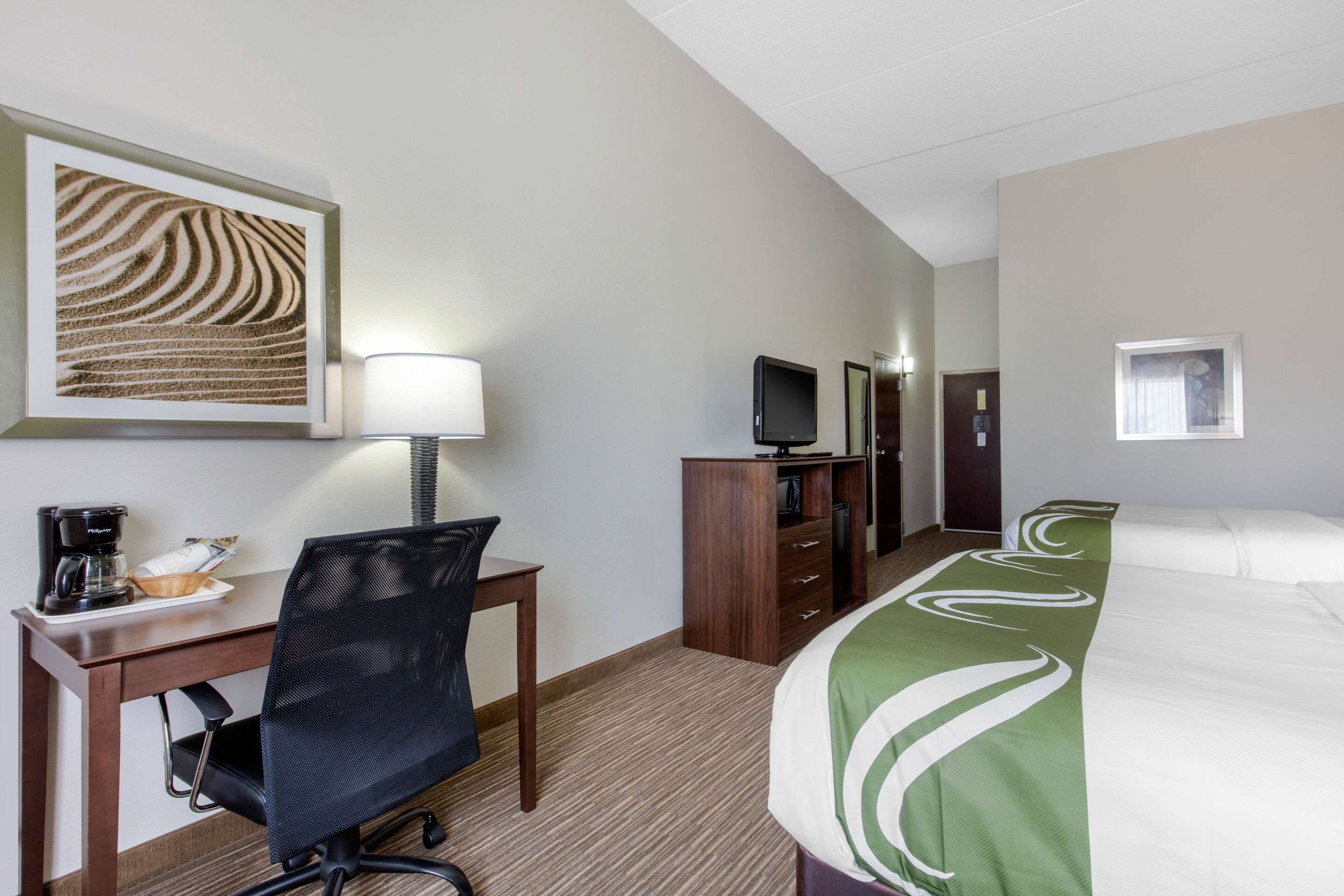 A hotel room with a bed, a desk with a chair, a television, a coffee maker, and a framed picture on the wall.