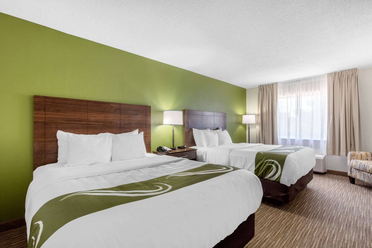 A hotel room with two beds, a green wall, a wooden headboard, a nightstand, a lamp, a chair, and a window with curtains.