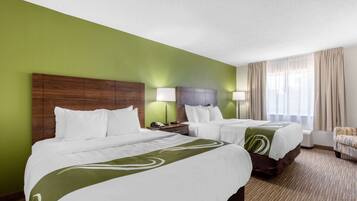 A hotel room with two beds, a green wall, a wooden headboard, a nightstand, a lamp, a chair, and a window with curtains.