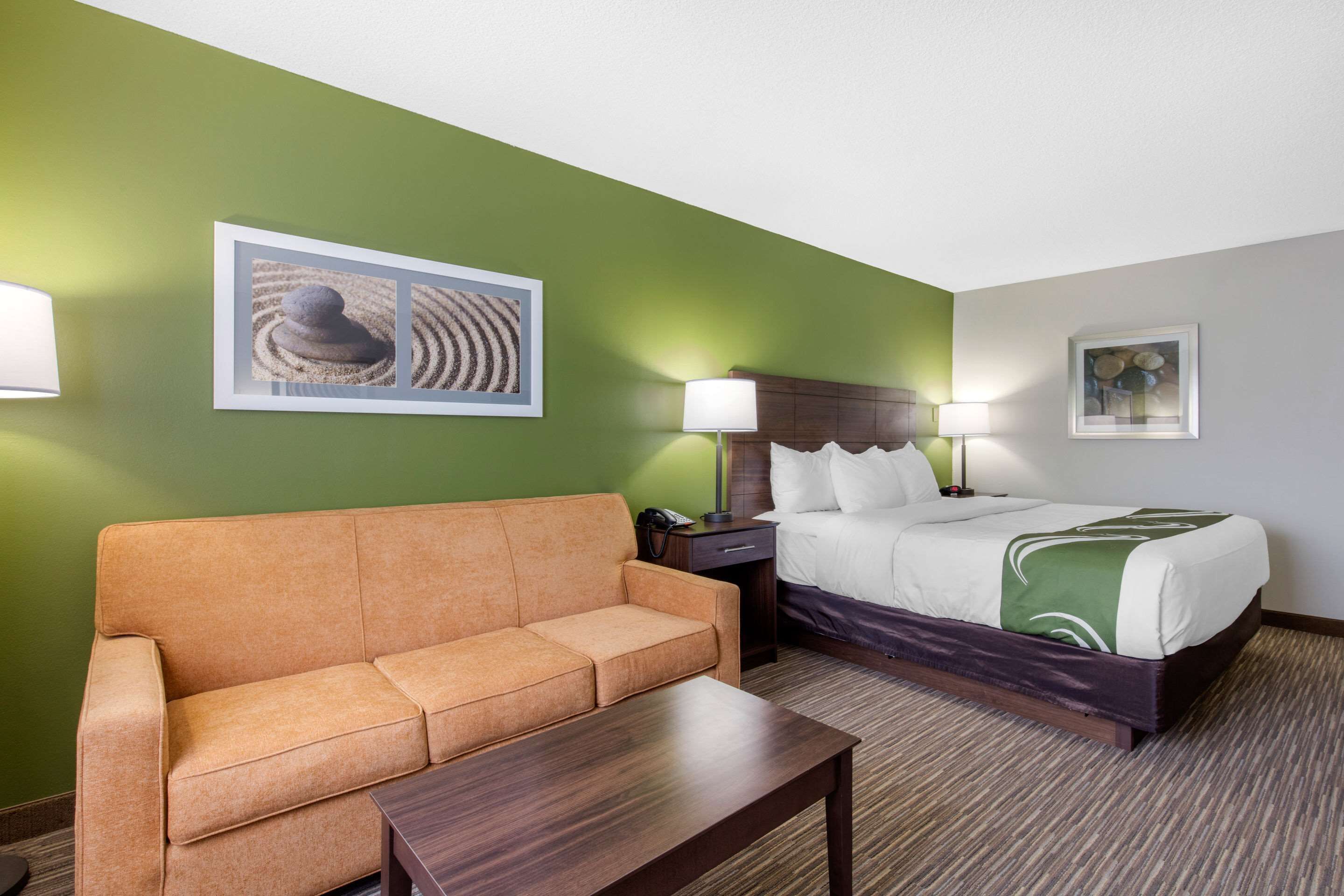 A hotel room with a green wall, a bed with white bedding, a brown sofa, a wooden nightstand, and a lamp.