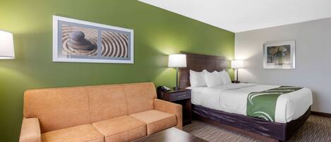 Habitación de hotel con pared verde, cama con ropa de cama blanca, sofá marrón, mesita de noche de madera y una lámpara.