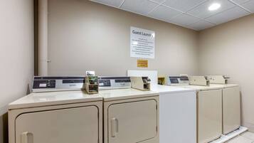 Une buanderie équipée de plusieurs machines à laver et de plusieurs séchoirs, un panneau indiquant que cet espace est réservé à la lessive des clients, et un panneau de commande pour les machines.