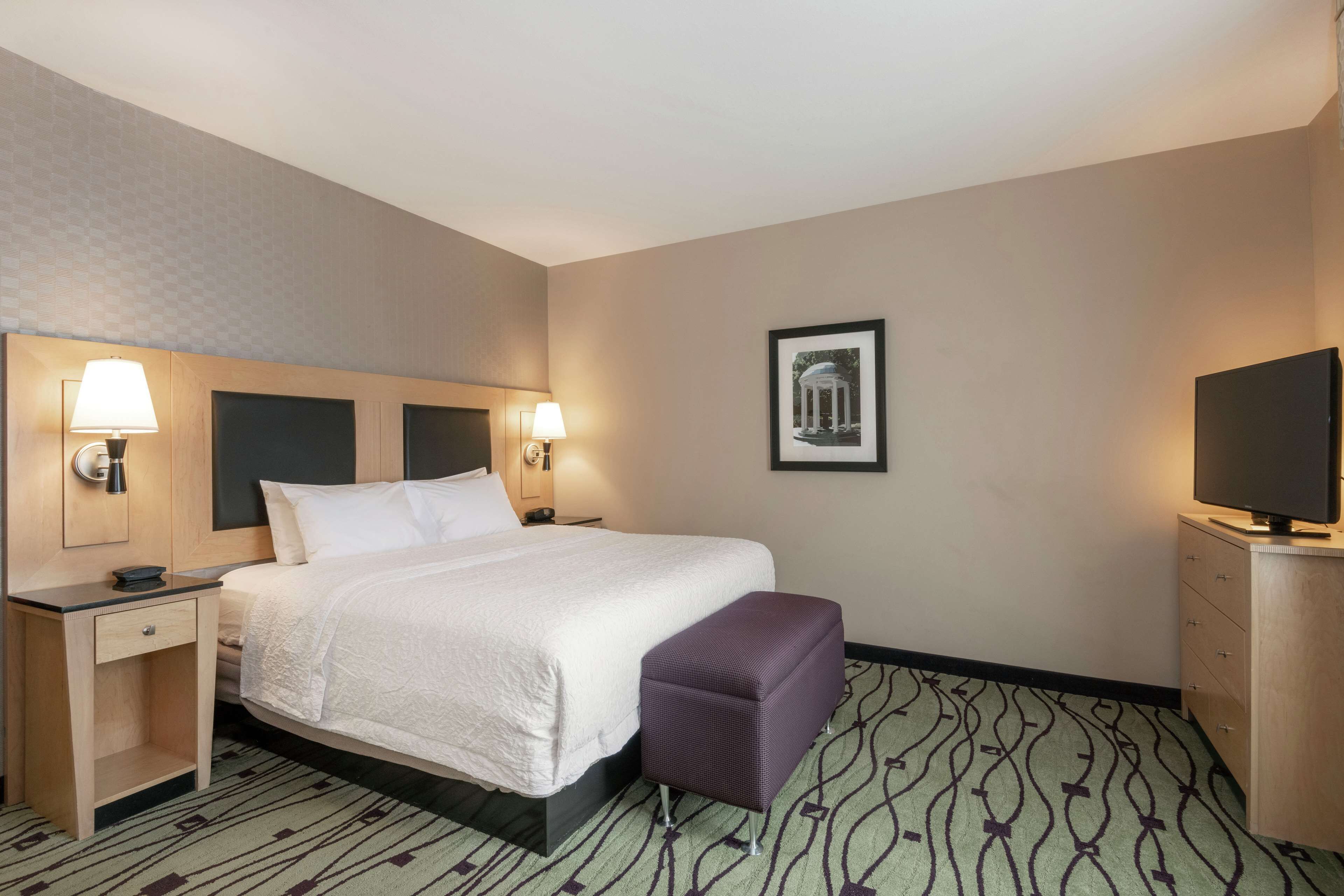 A hotel room with a bed, bedside tables, a desk, a television, and a patterned carpet.