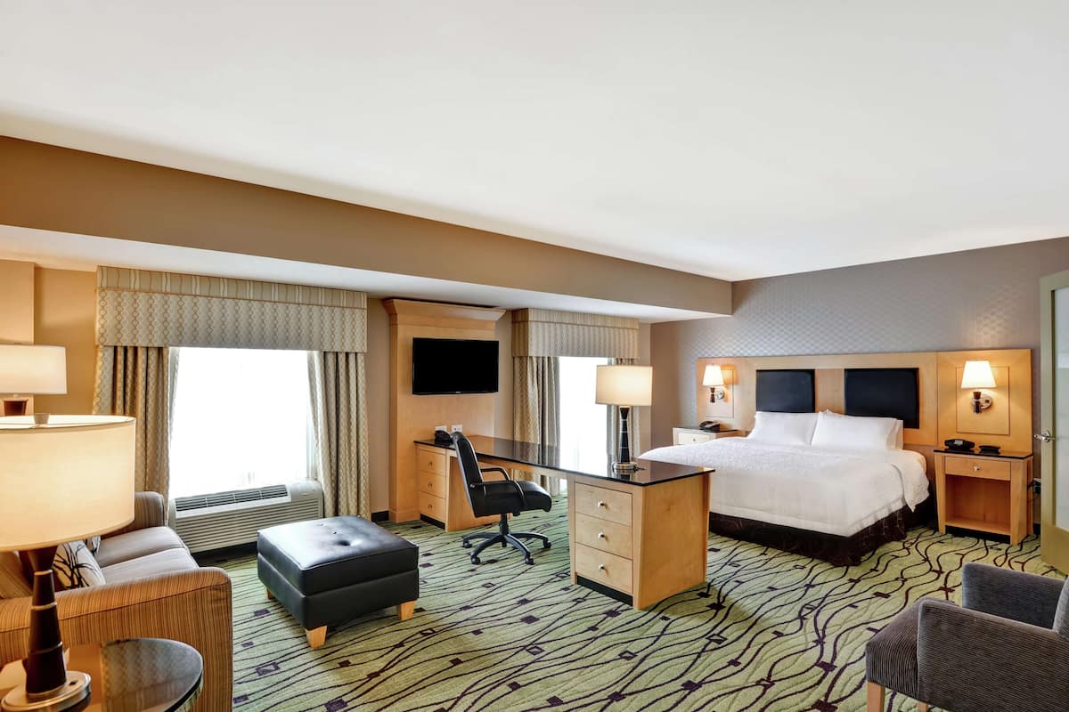 A hotel room with a large bed, a desk, a chair, a sofa, a TV, and a lamp.