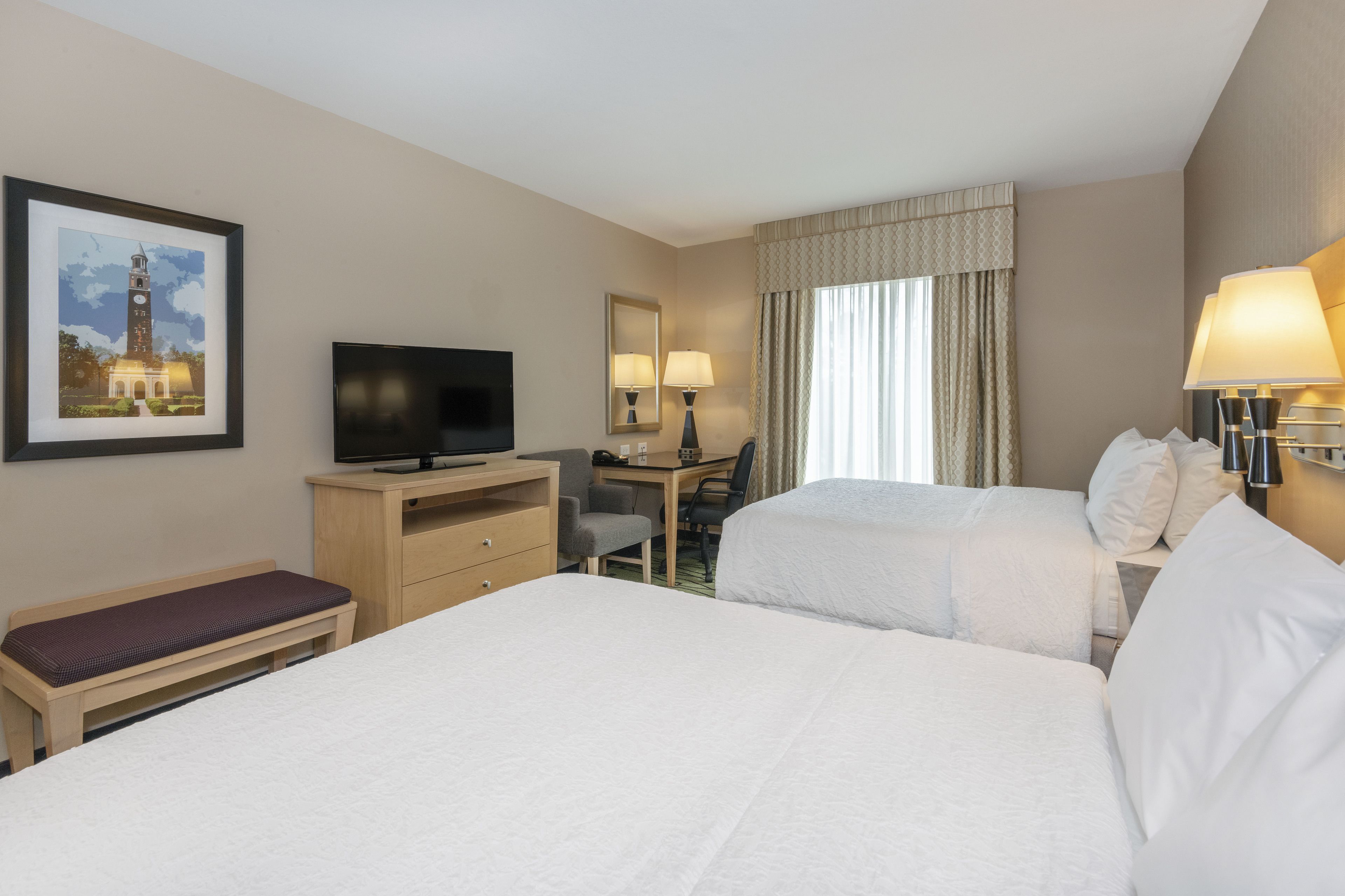 A hotel room with two beds, a desk, a television, a bench, and a framed picture on the wall.