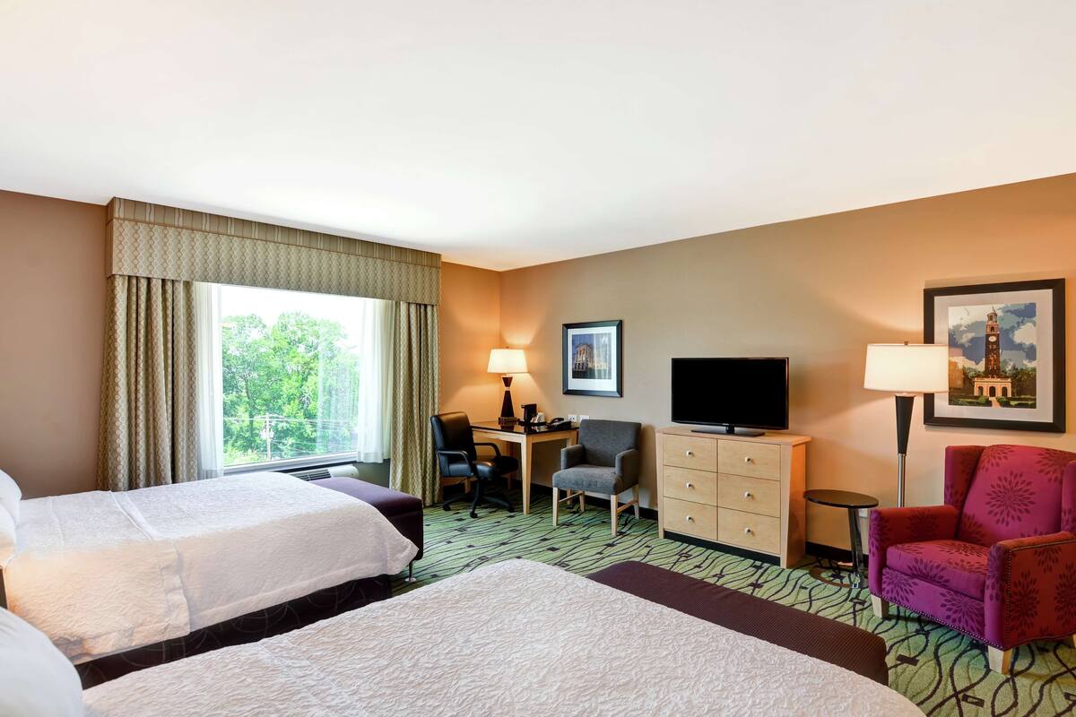 A hotel room with two beds, a desk, a television, a chair, and a lamp.