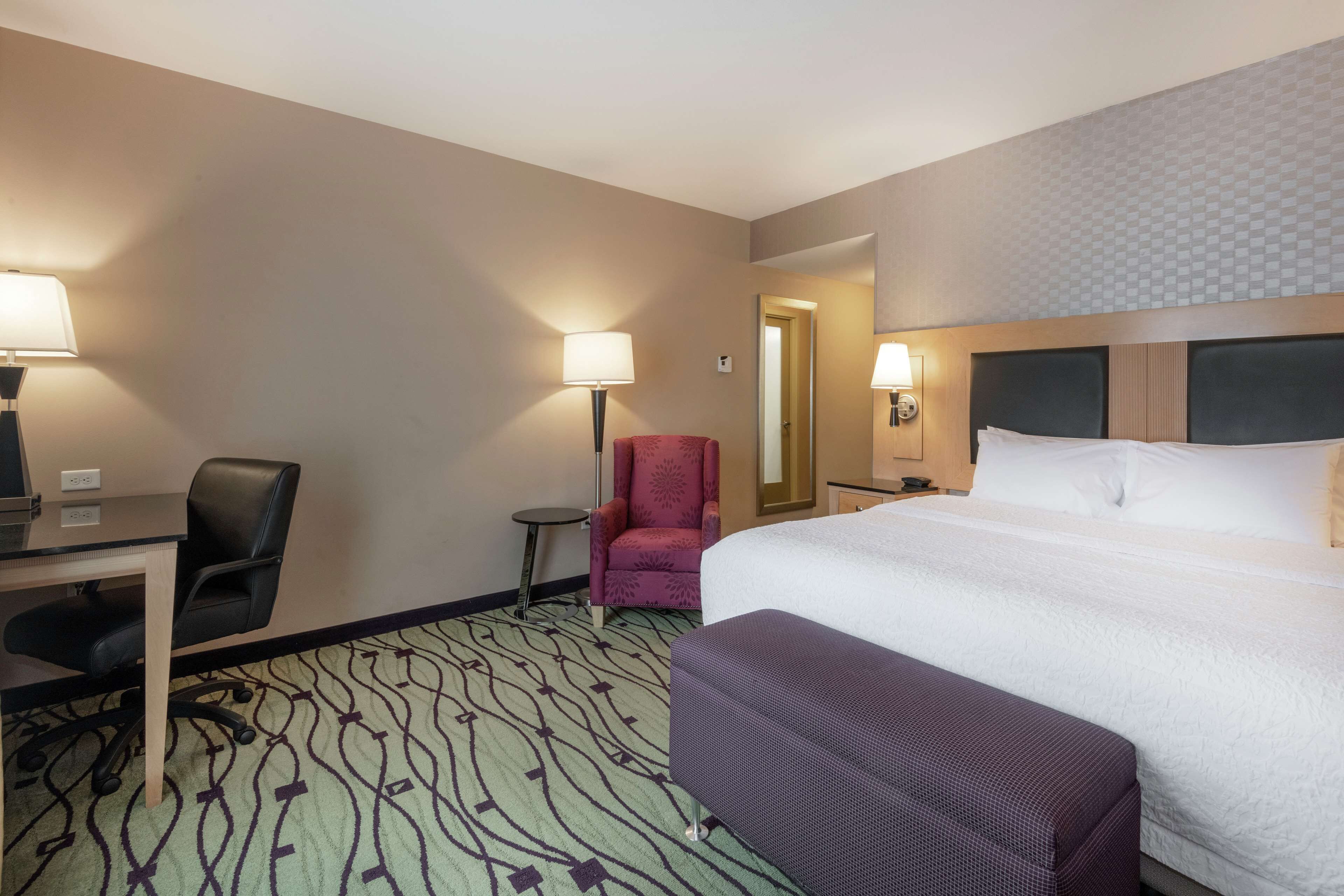 A hotel room with a bed, a desk, a chair, a small table, and a lamp.