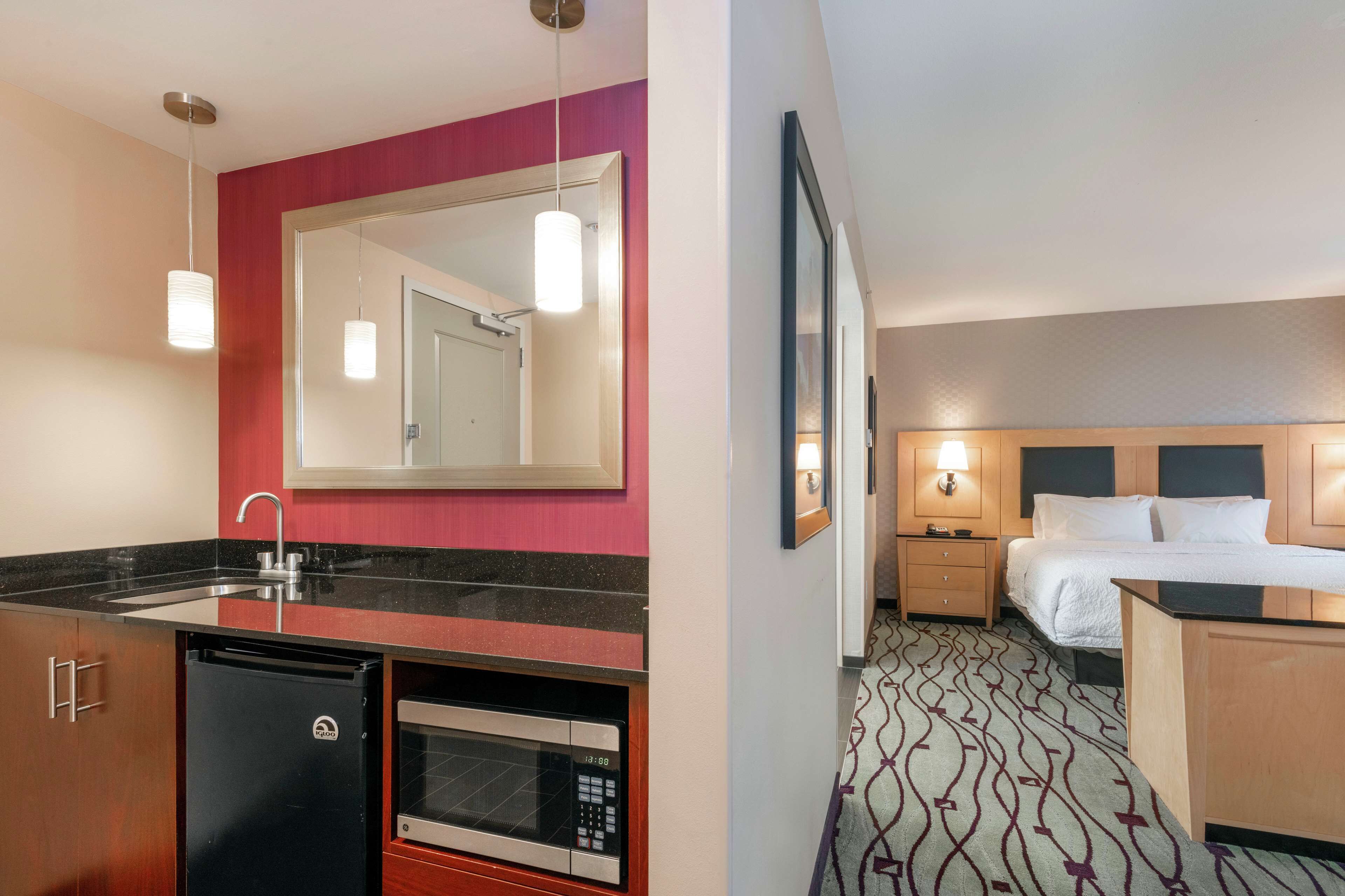 A hotel room with a bed, bedside table, and a small desk. There is a microwave and a mini-fridge in the kitchenette area.