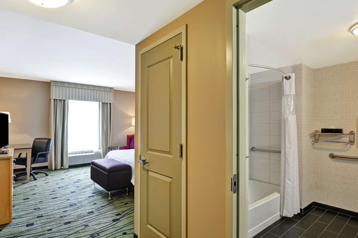 A hotel room with a bed, a desk, a chair, a bathroom with a shower, and a window with curtains.