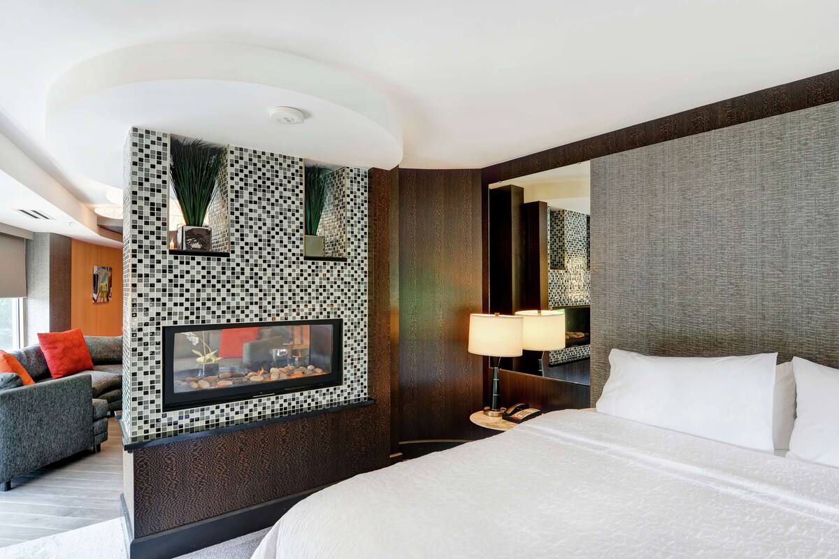 A hotel room with a large bed, a TV mounted on a mosaic wall, a nightstand with a lamp, and a sofa with orange cushions.