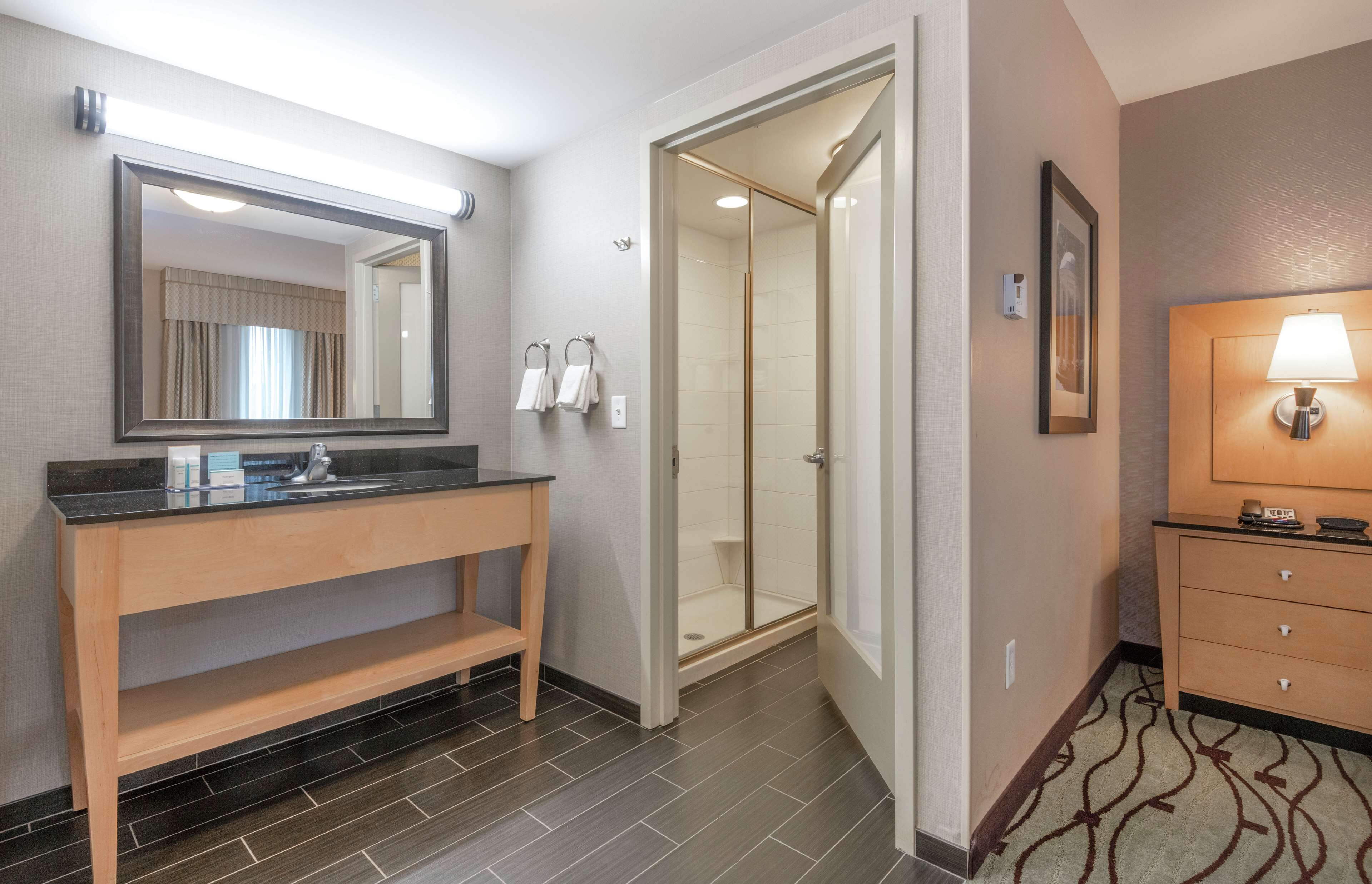 A modern bathroom with a vanity, mirror, shower, and a bedside lamp.
