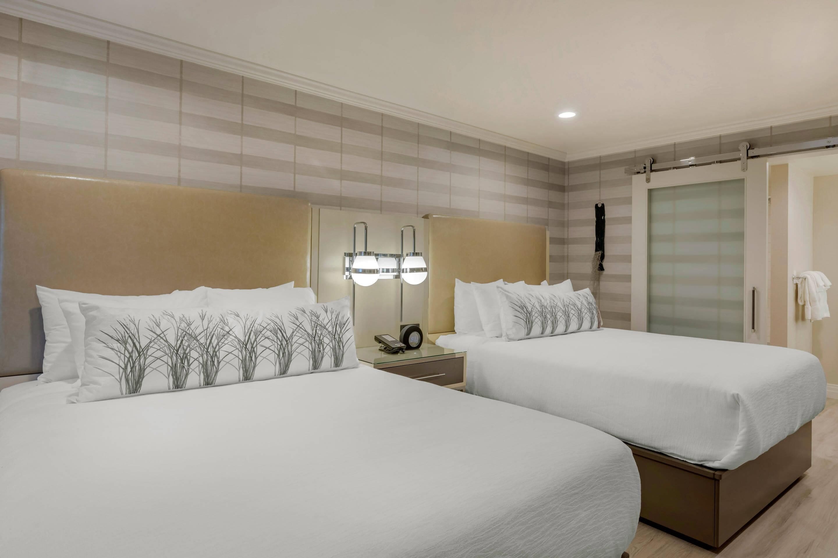 Habitación de hotel con dos camas, una mesita de noche con lámpara, un espejo y un armario con puerta corredera.