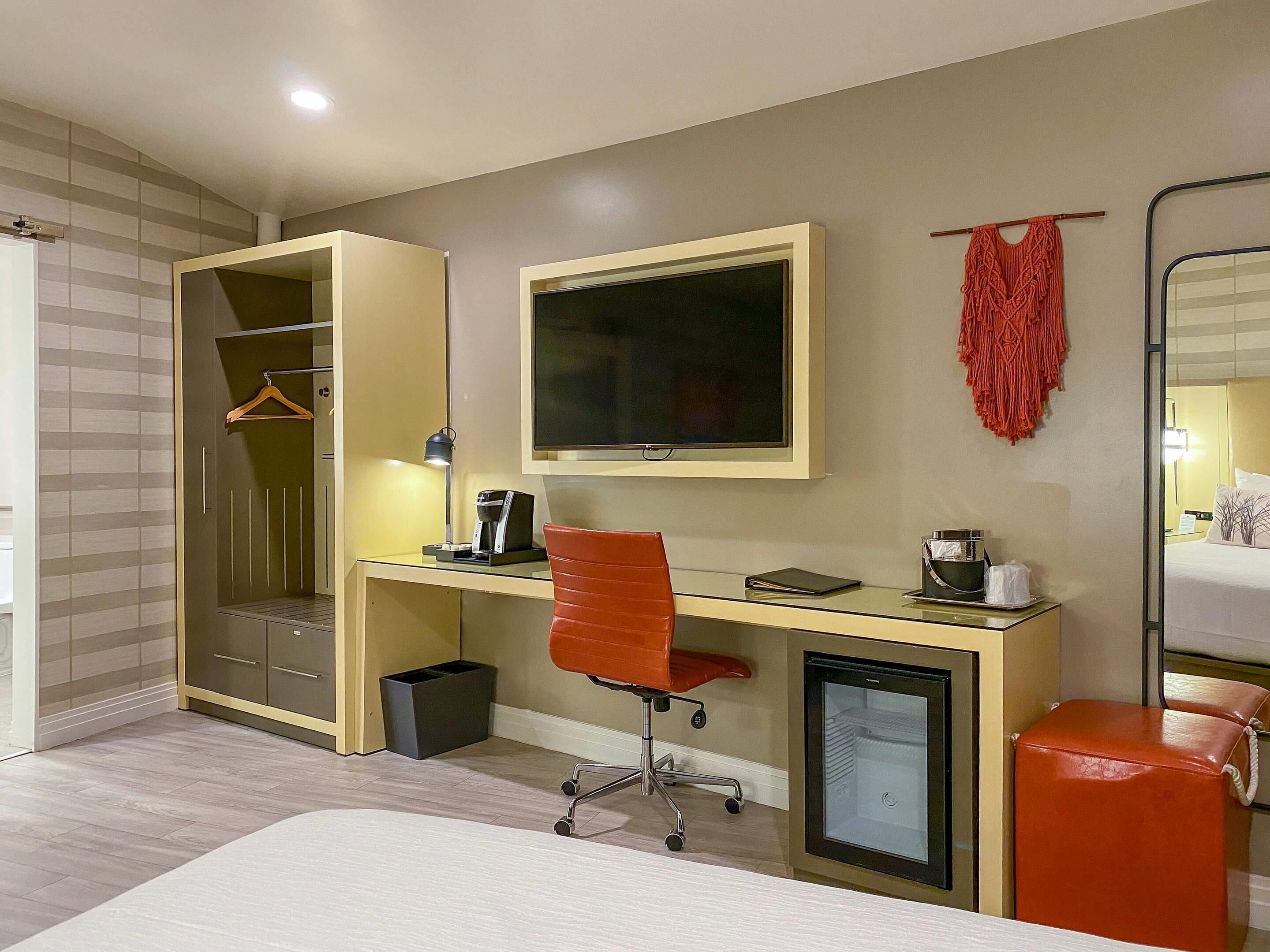 A hotel room with a bed, a desk with a computer, a chair, a TV, a small fridge, and a wardrobe.