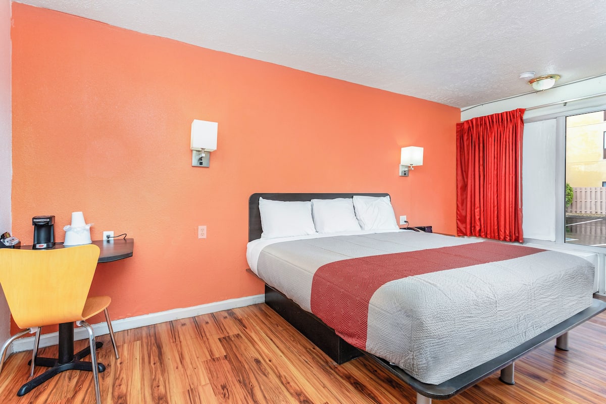 A hotel room with a bed, a desk with a chair, and a window with red curtains.