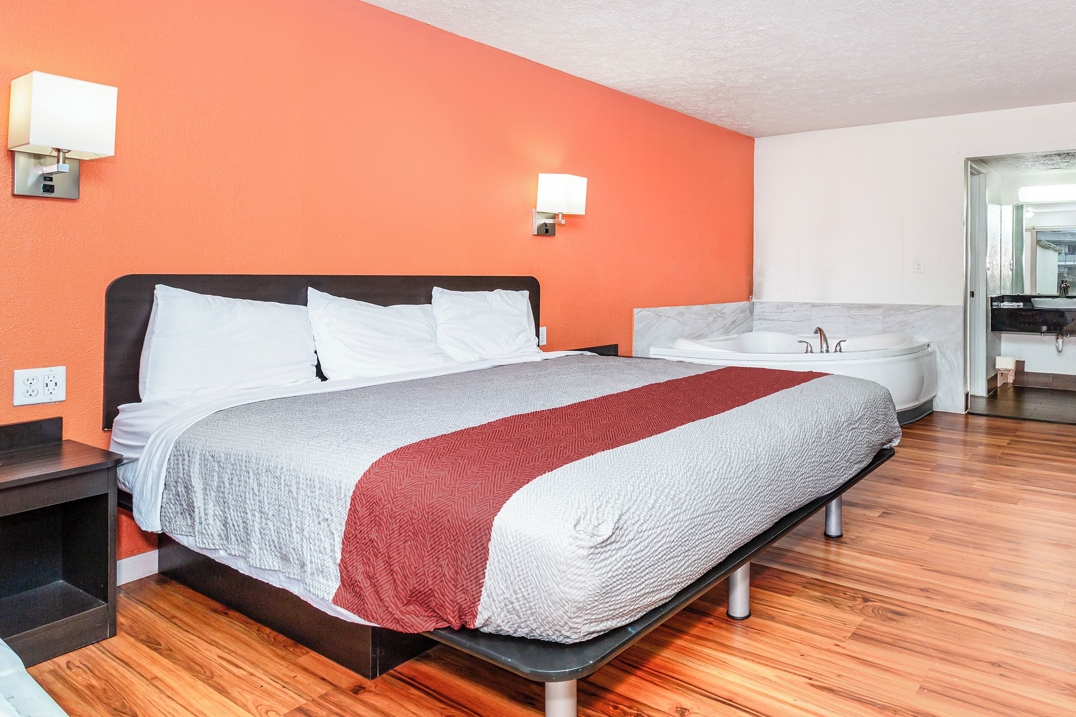 Quarto de hotel com cama, mesa de cabeceira, banheiro com pia e espelho, e uma parede laranja.