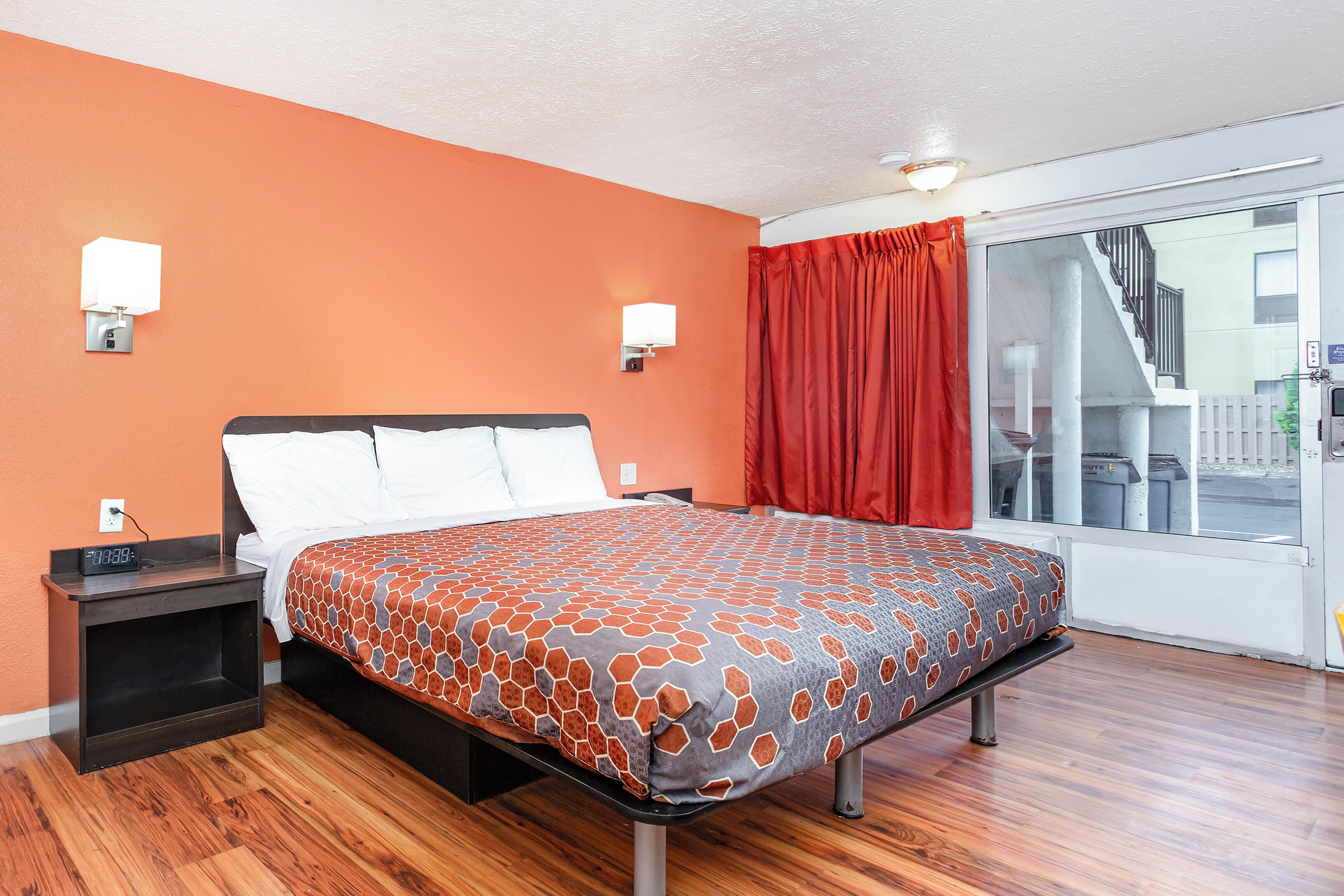 Una habitación de hotel con cama, suelo de madera, mesita de noche, ventana con vistas al exterior y dos luces fijadas a la pared.