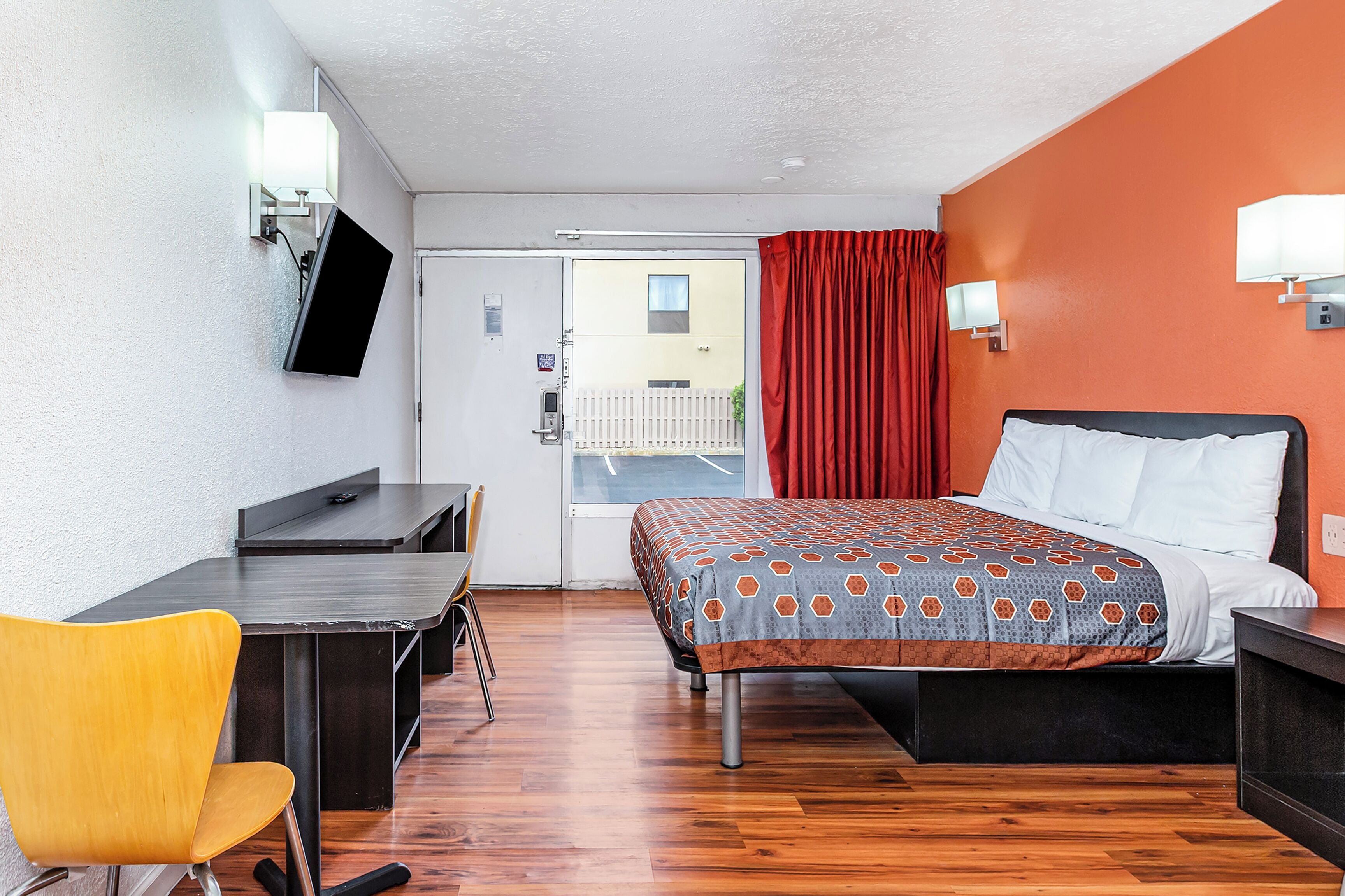 A hotel room with a bed, a desk, a chair, a TV mounted on the wall, and a sliding door to the outside.