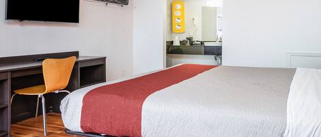 A hotel room with a bed, a desk, a chair, a TV, and a bathroom visible through an open door.