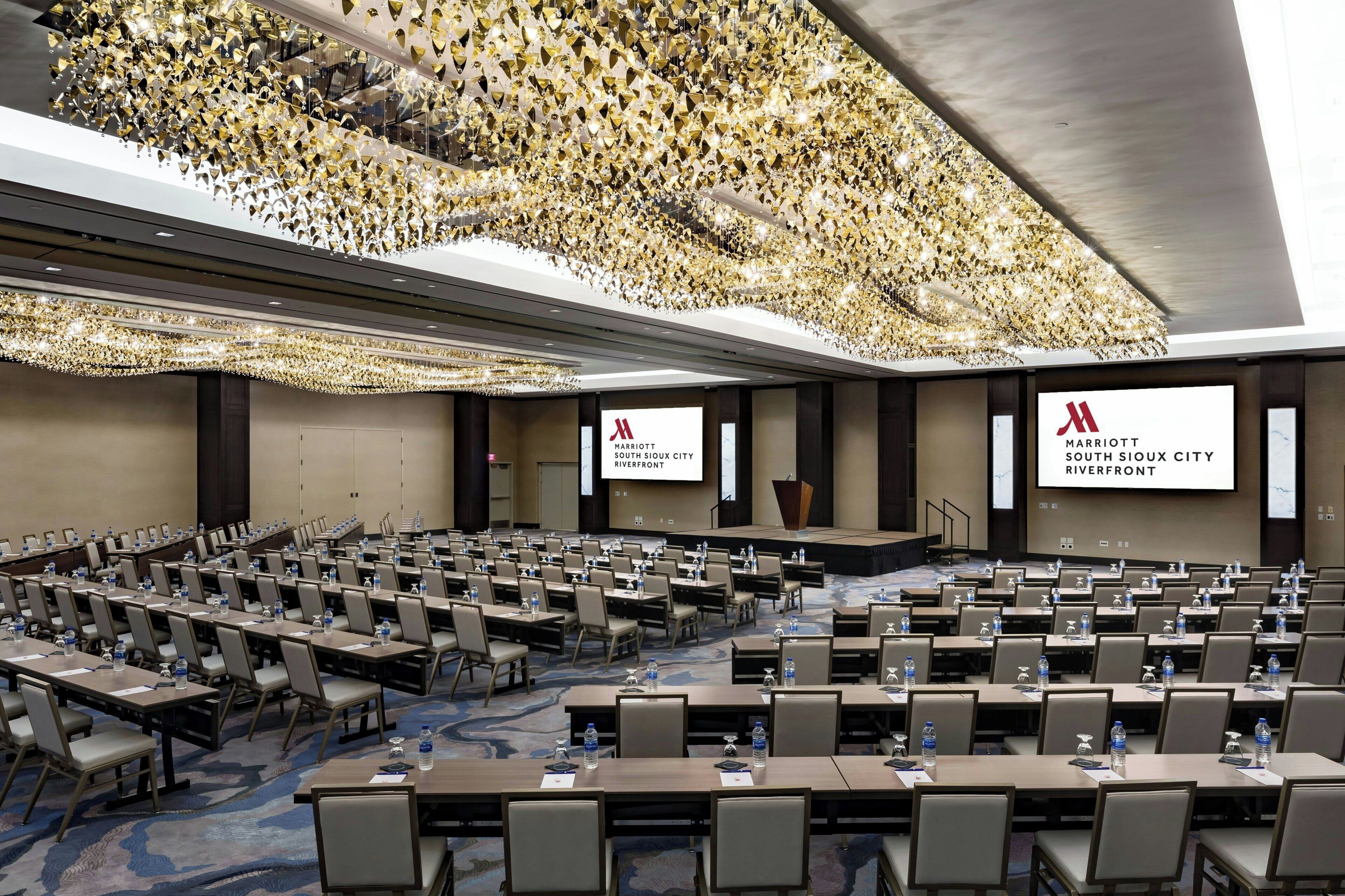 Un amplio salón de conferencias con filas de mesas y sillas, una pantalla grande mostrando el logotipo de Marriott, y un techo decorativo.