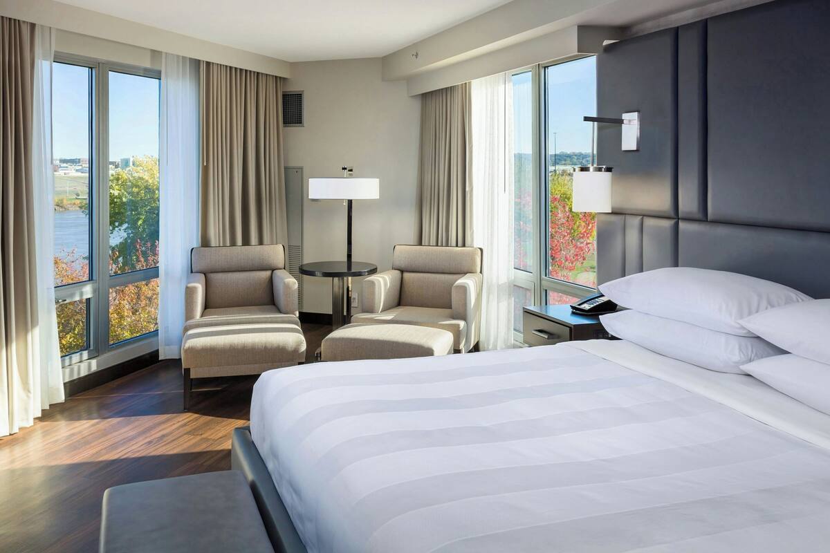 Ein Hotelzimmer mit einem großen Bett, zwei Sesseln, einem kleinen Tisch und Blick auf ein Gewässer durch große Fenster.