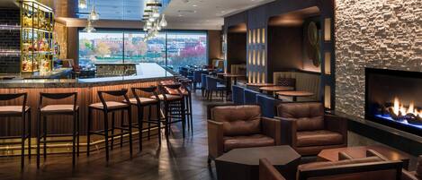 A bar area with stools and a fireplace, with a view of the cityscape through large windows.
