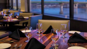 Mesa de comedor dispuesta con copas de vino, platos y cubiertos, con vistas a un puente y al agua a través de amplias ventanas.