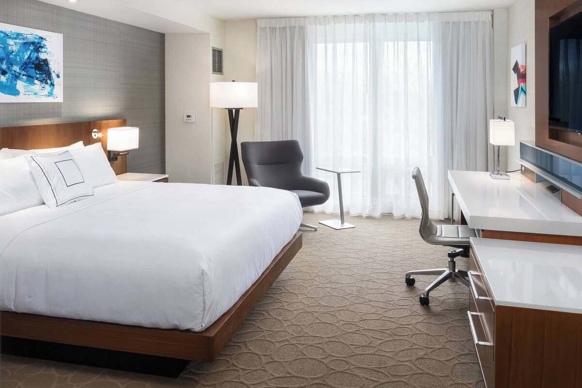 Ein Hotelzimmer mit einem großen Bett, einem Schreibtisch mit Stuhl, einem Fernseher und einem Fenster mit Vorhängen.