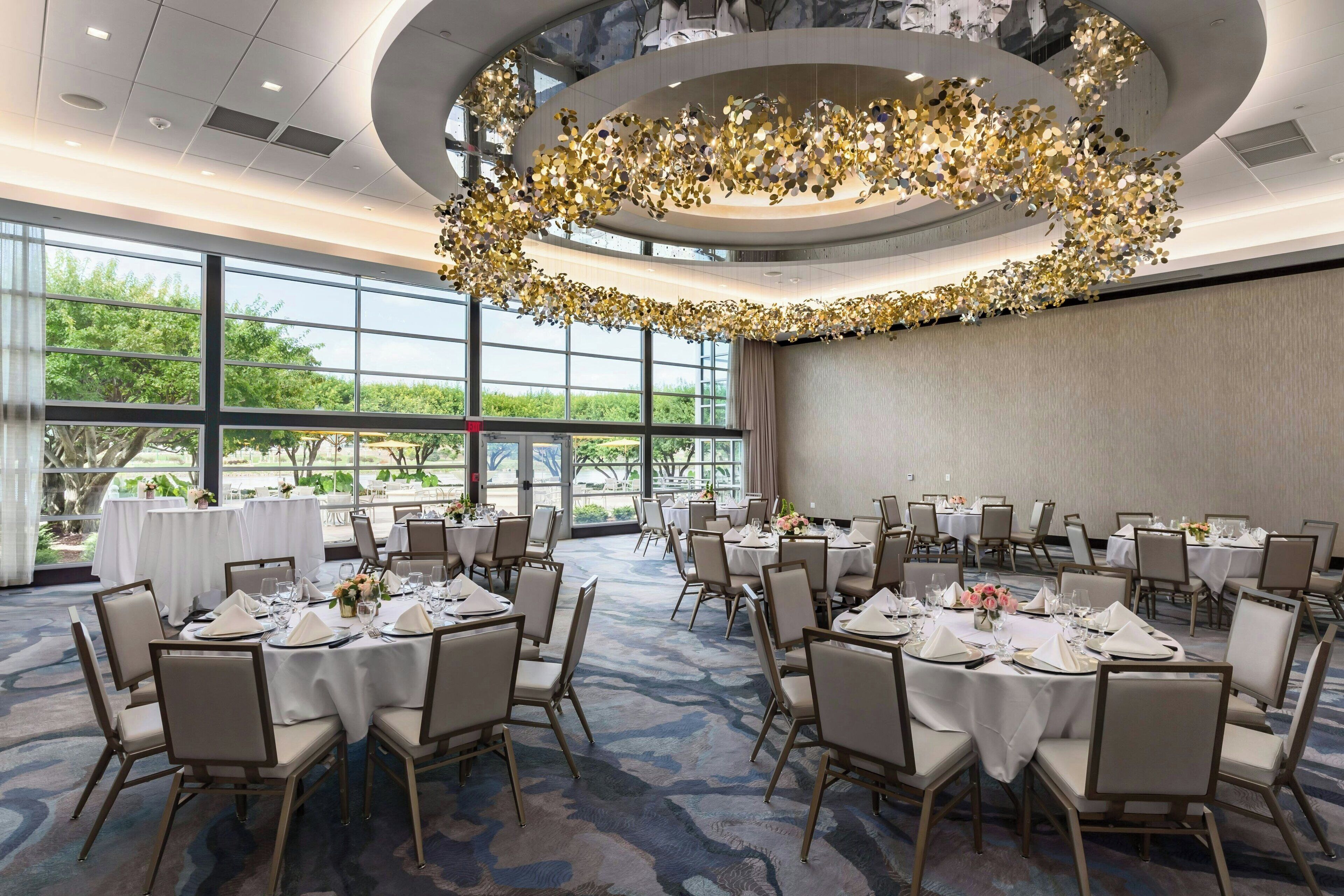 Un salón de banquetes con mesas redondas dispuestas para comer, grandes ventanales que ofrecen vistas a la vegetación y un elemento decorativo en el techo.