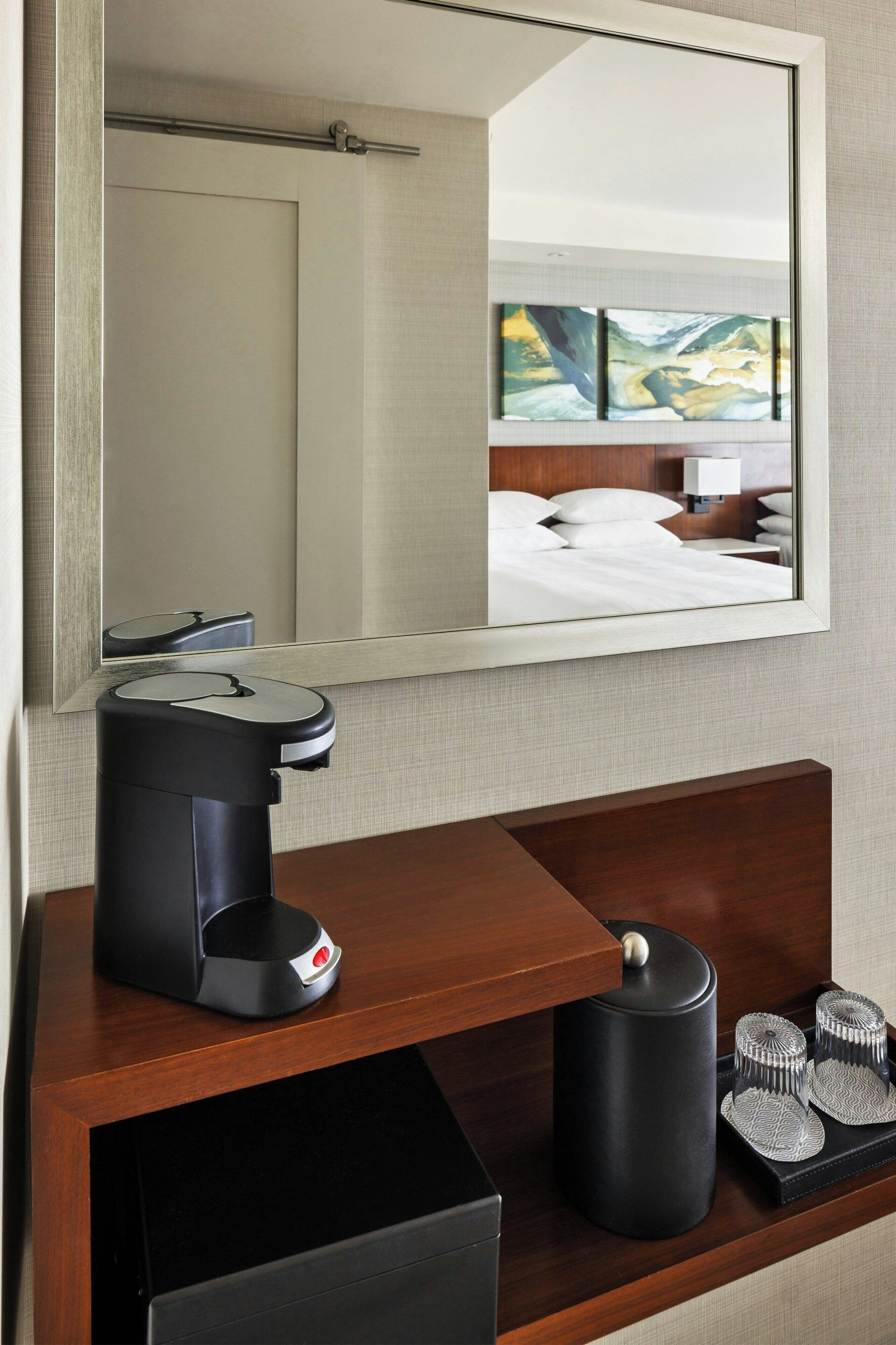 Ein Hotelzimmer mit einem Holzkonsolentisch, einer Kaffeemaschine, einem Mülleimer und einem Spiegel, der ein Bett und ein Gemälde reflektiert.