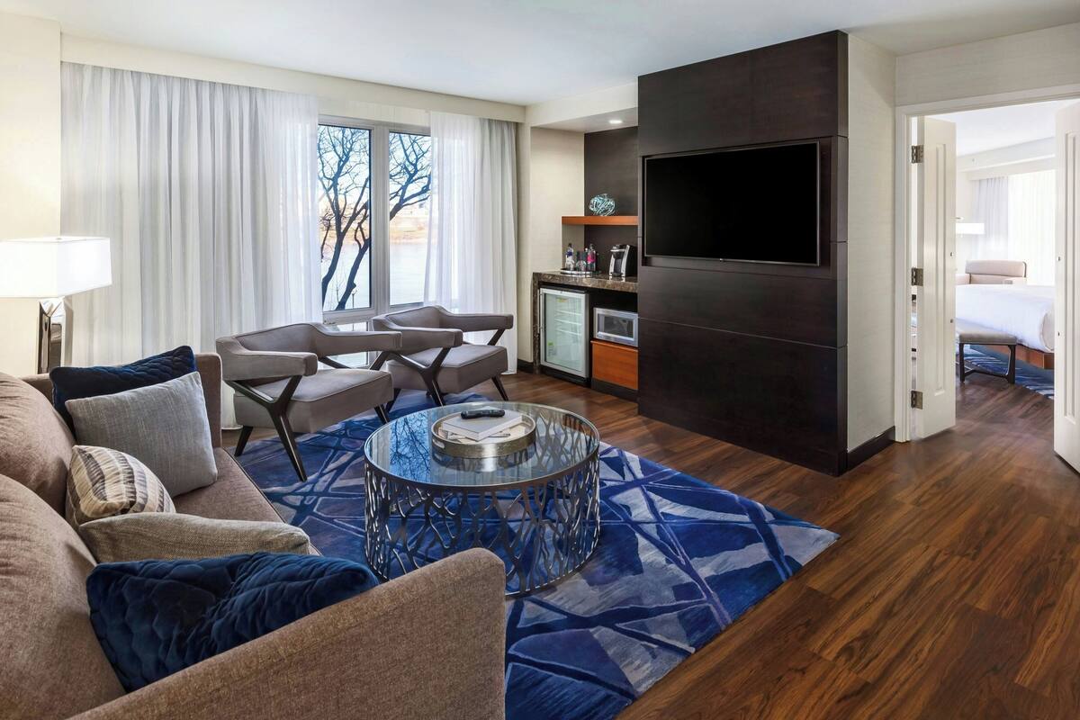 Ein modernes Hotelzimmer mit Wohnbereich, ausgestattet mit einer Couch, Sesseln, einem Glastisch und einem Flachbildfernseher, der an einer dunklen Wand angebracht ist.