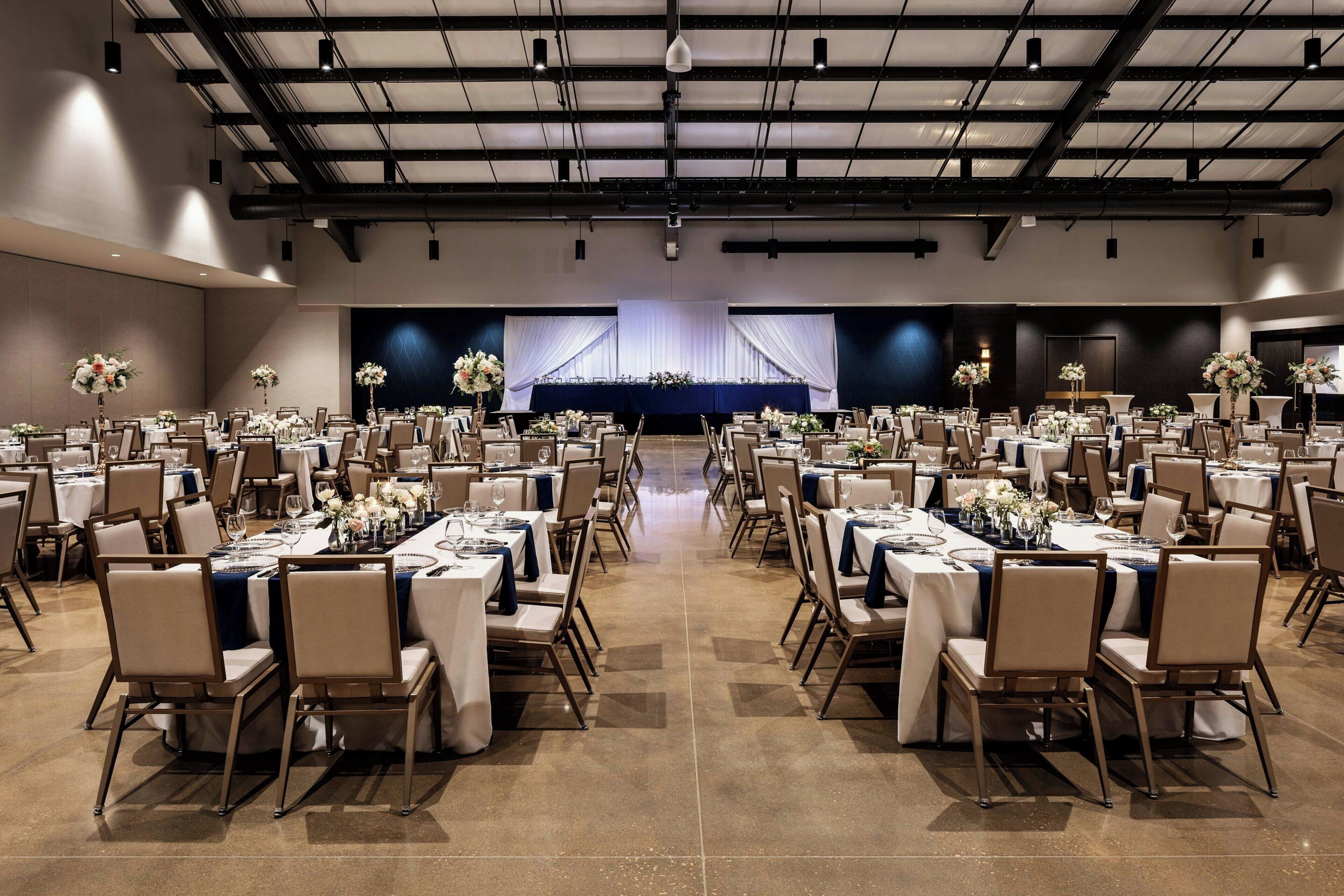 A large banquet hall with rows of tables and chairs, a stage with draped curtains, and a high ceiling with exposed beams.