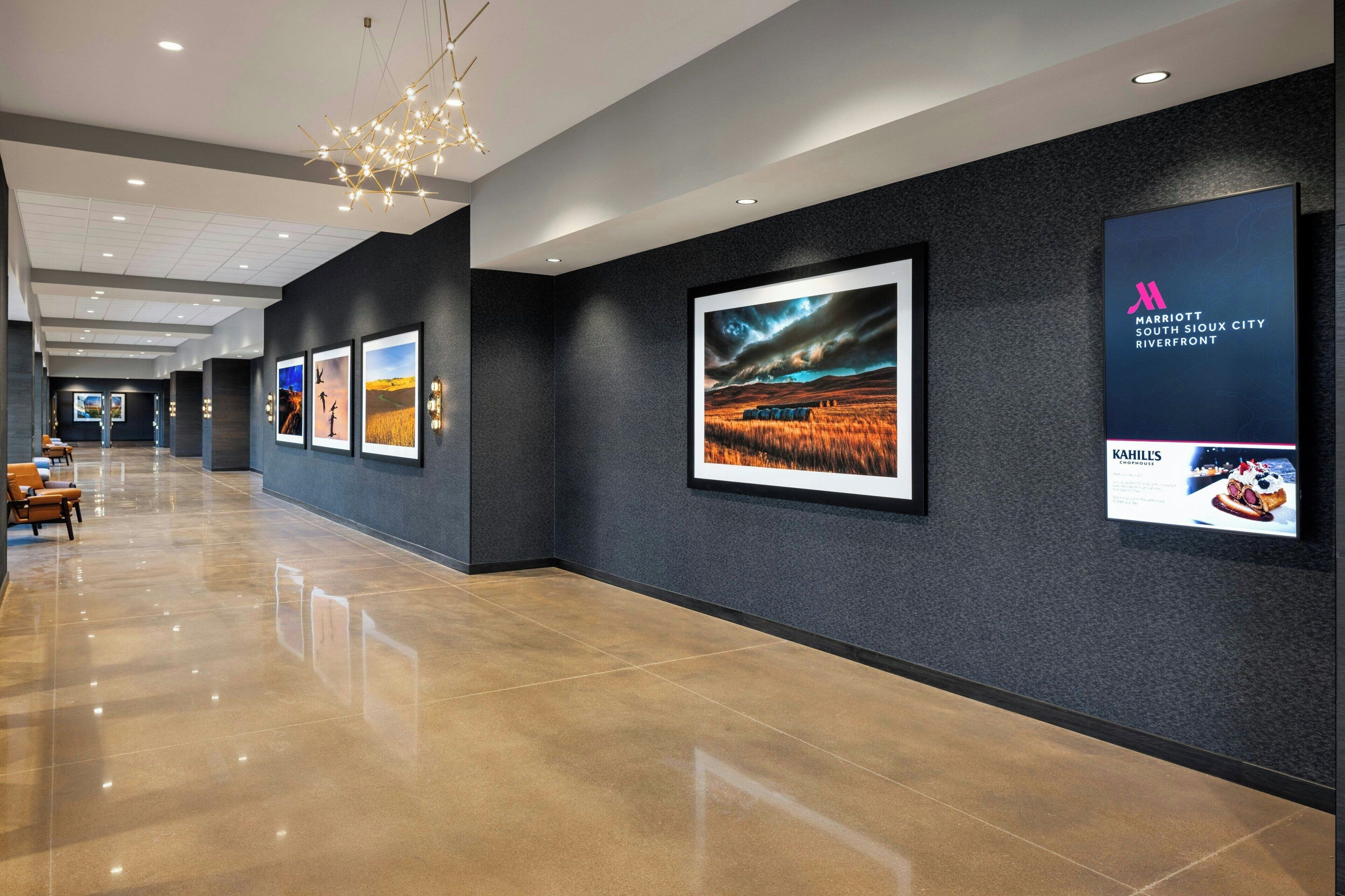 A modern hotel corridor with framed artwork on the walls, a polished floor, and a chandelier.