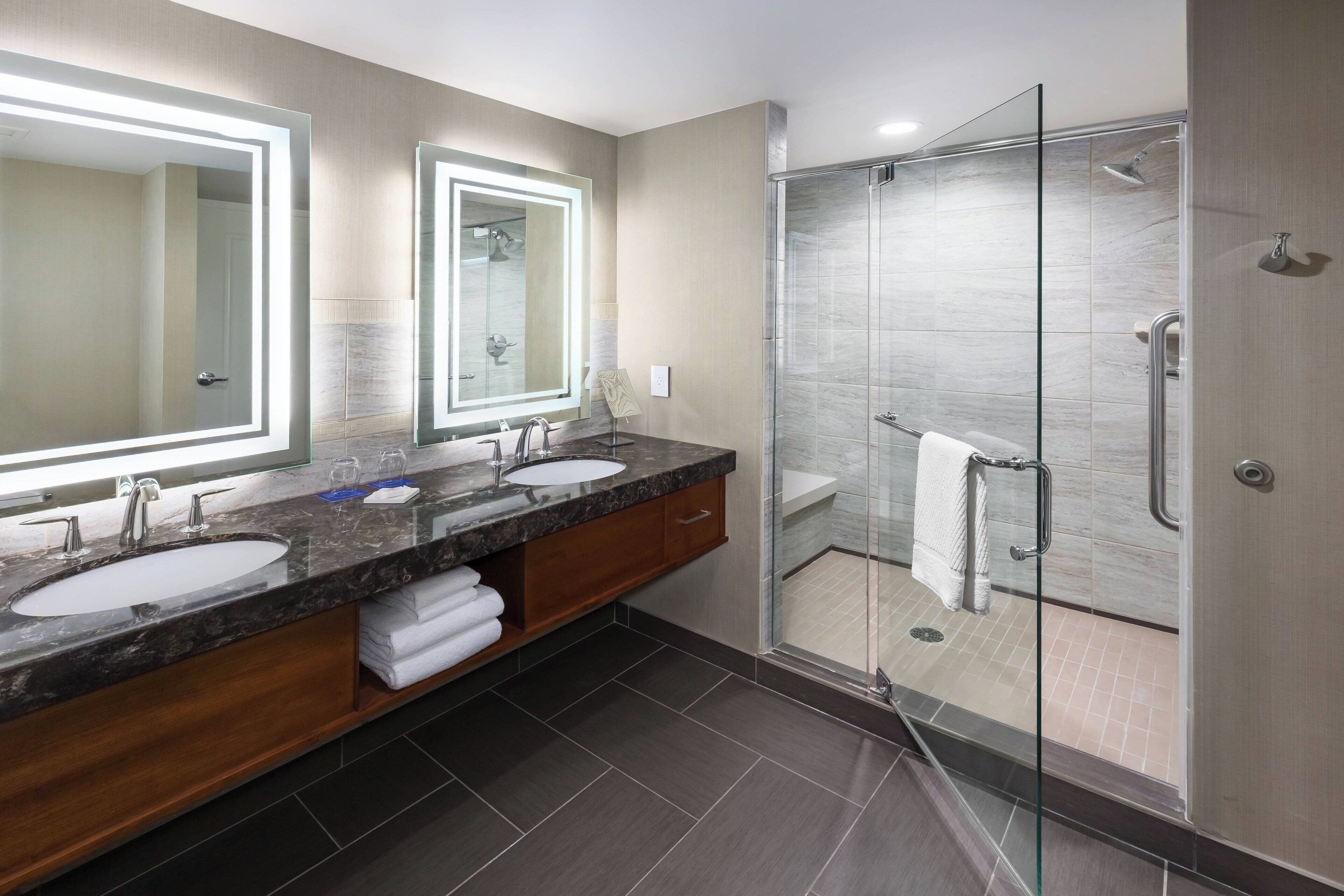 A modern bathroom with two sinks, a glass-enclosed shower, and a large mirror with under-cabinet lighting.