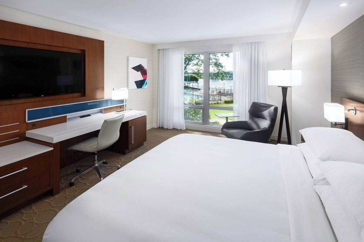 Ein Hotelzimmer mit einem großen Bett, einem Schreibtisch mit Stuhl, einem Flachbildfernseher, einer Lampe und Blick auf Pool und Grünflächen.