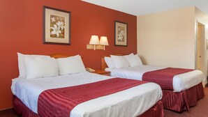 A hotel room with two beds, each with white bedding and red skirts, a wooden headboard, a nightstand with a lamp, and two framed floral pictures on the wall.