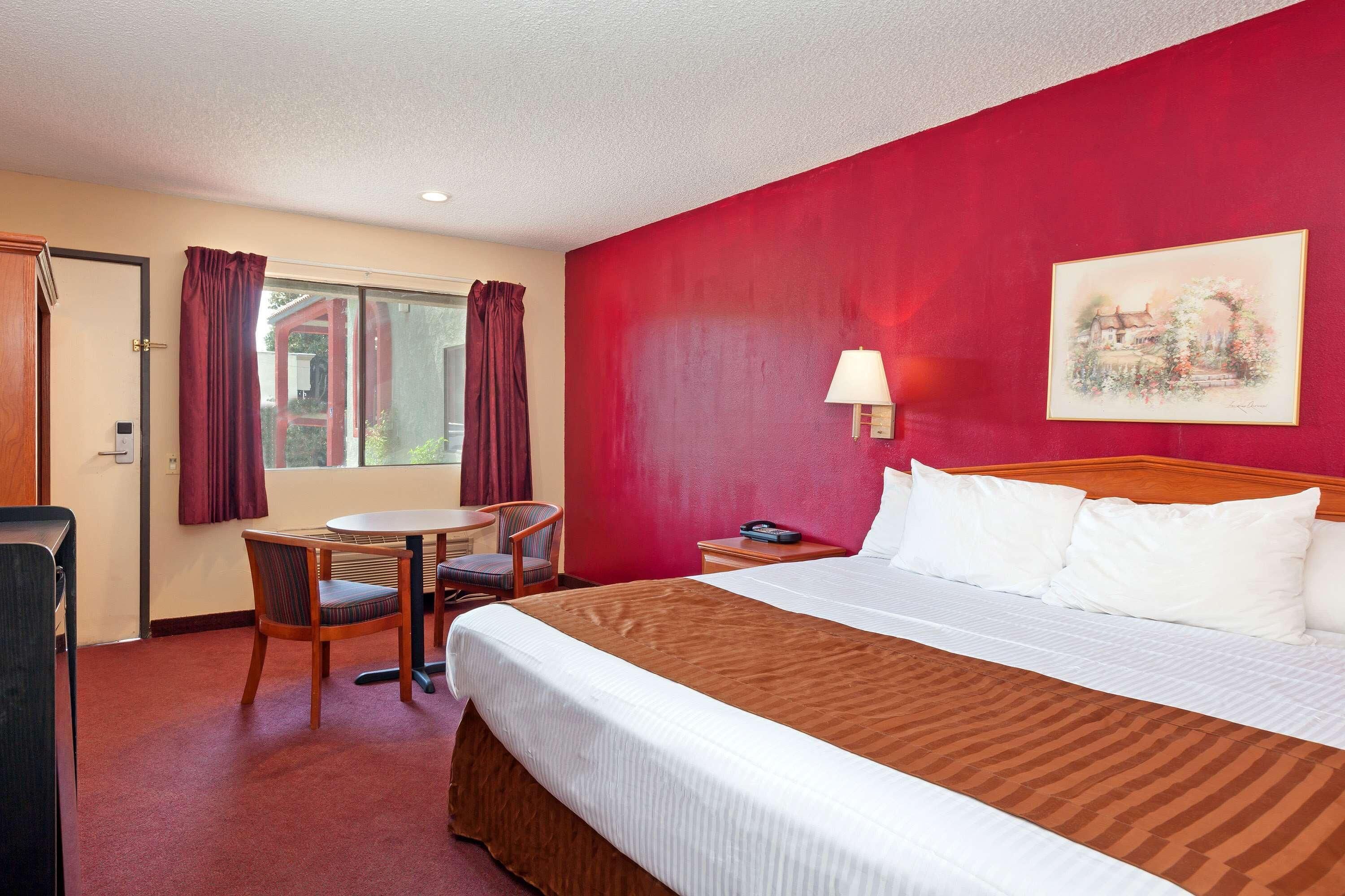 Une chambre d’hôtel comprenant un lit, un bureau, une chaise, une fenêtre avec des rideaux, une table de chevet et un tableau au mur.