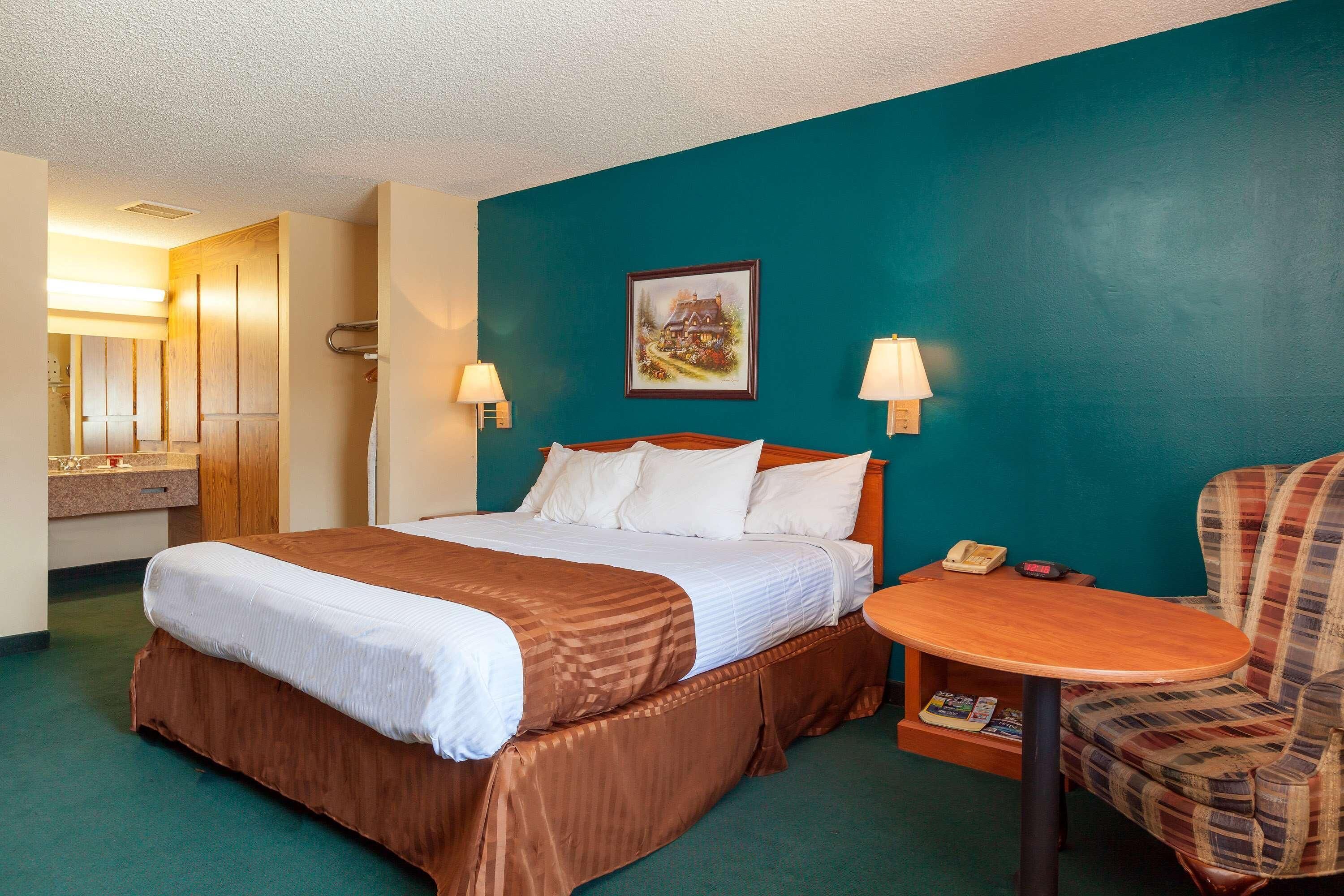 Une chambre d’hôtel avec un lit, une petite table, une chaise, un tableau accroché au mur et une salle de bain visible à travers une porte ouverte.