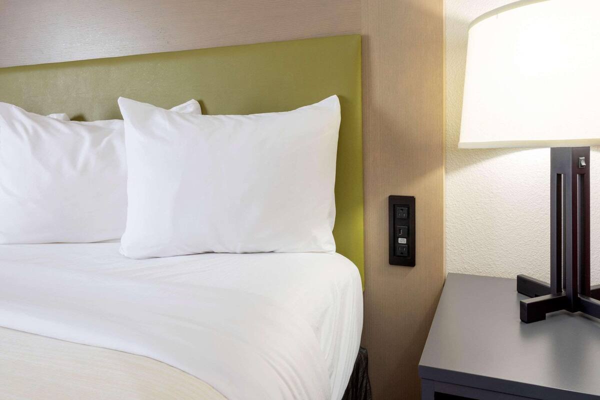 A hotel room with a bed, white pillows, a green headboard, a bedside table with a lamp, and a control panel on the wall.