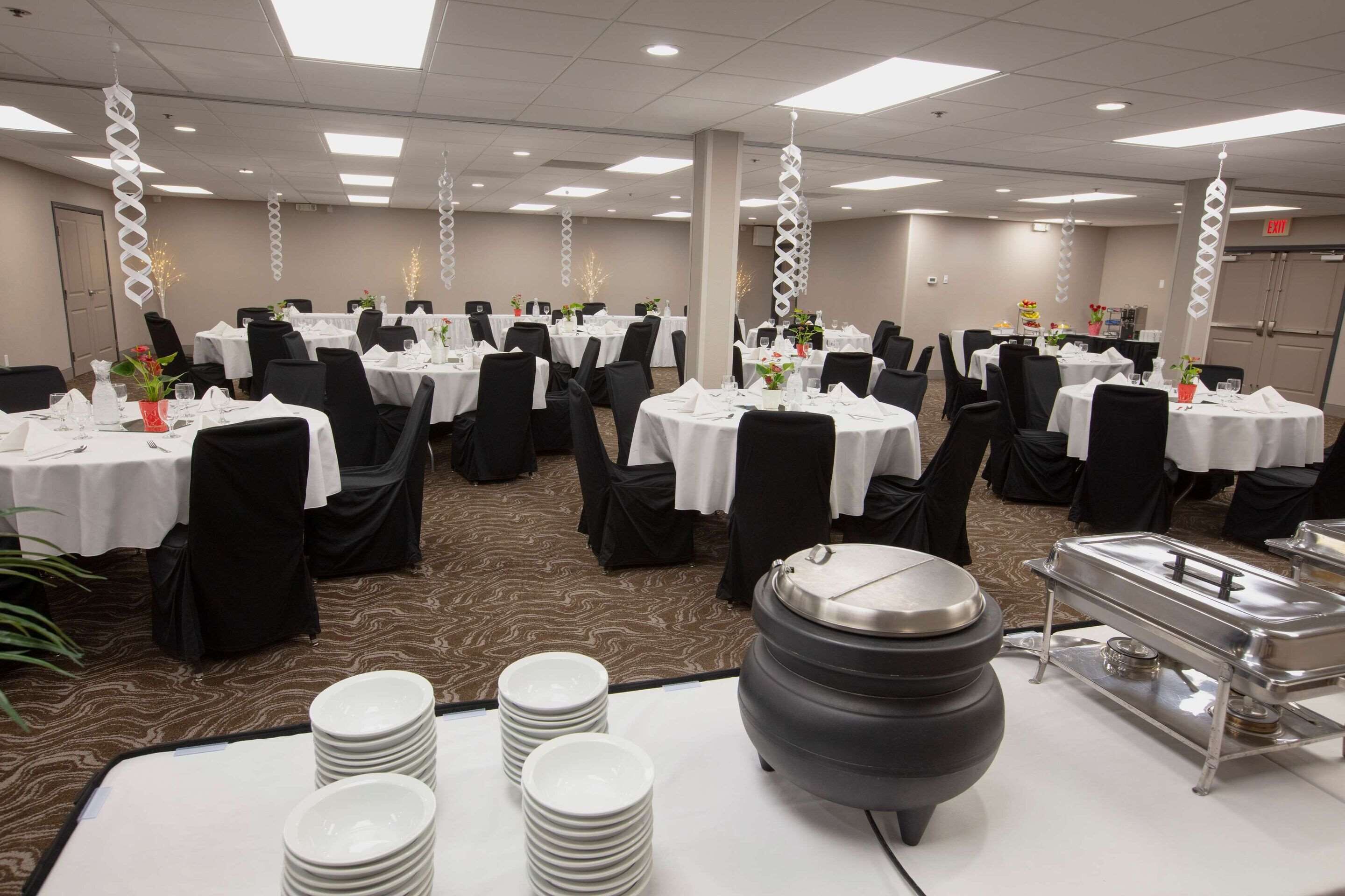 A banquet hall with round tables, black chairs, white tablecloths, and a buffet station.